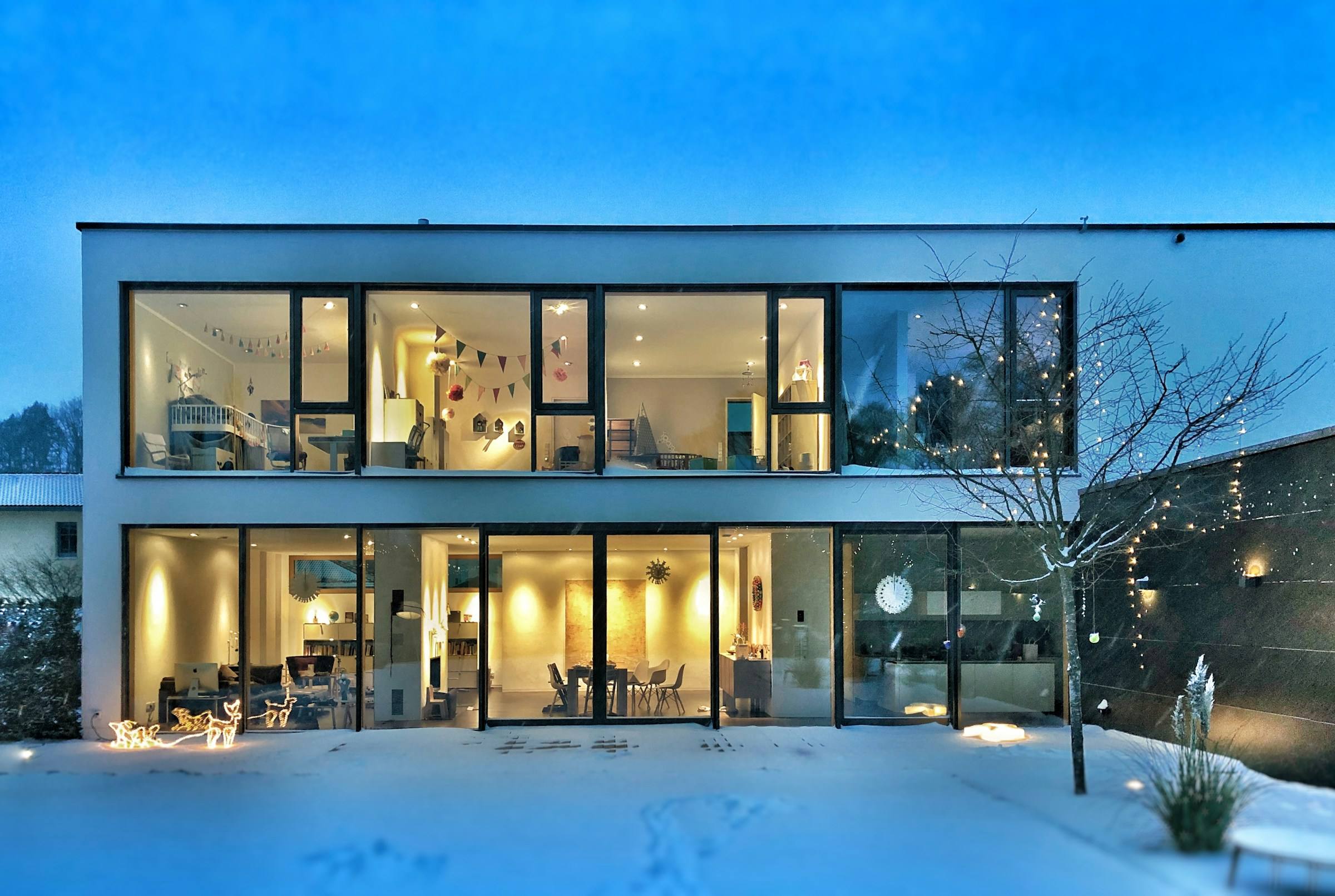 Modern two-story house at dusk, its floor-to-ceiling windows glowing warm against the snow